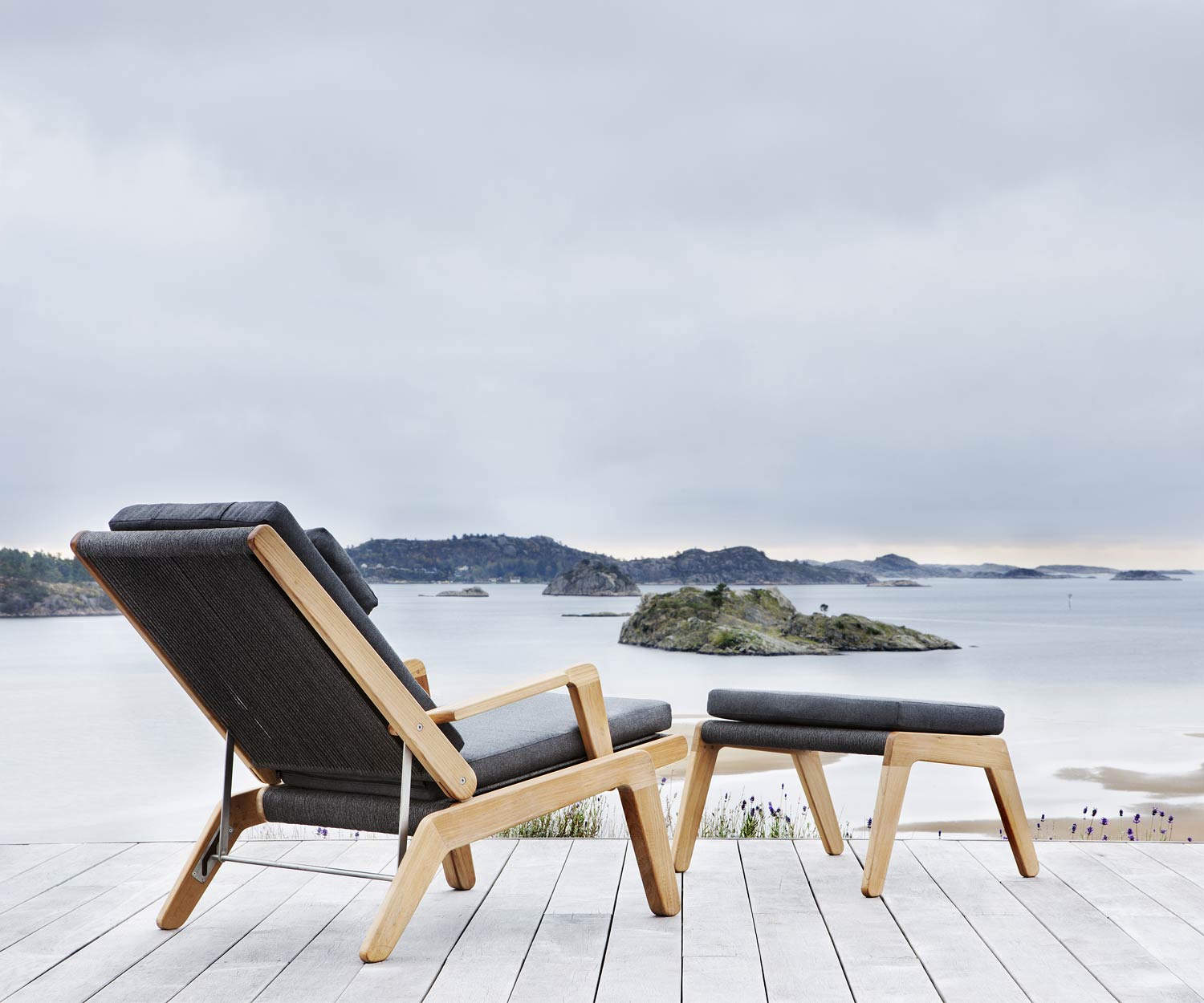 Oasiq Sgabello di design Skagen con cuscino imbottito in grigio scuro davanti alla sedia a sdraio Oasiq Sgabello di design Skagen con cuscino imbottito in grigio scuro davanti alla sedia a sdraio